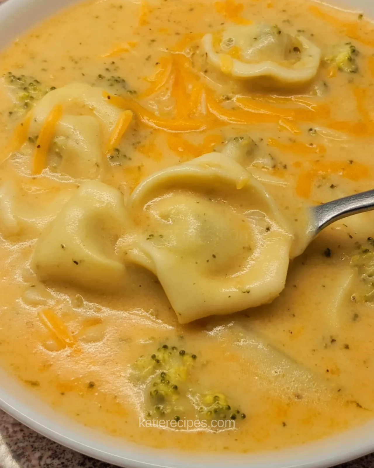 Creamy broccoli cheddar tortellini soup with broccoli florets and shredded cheese in a white bowl