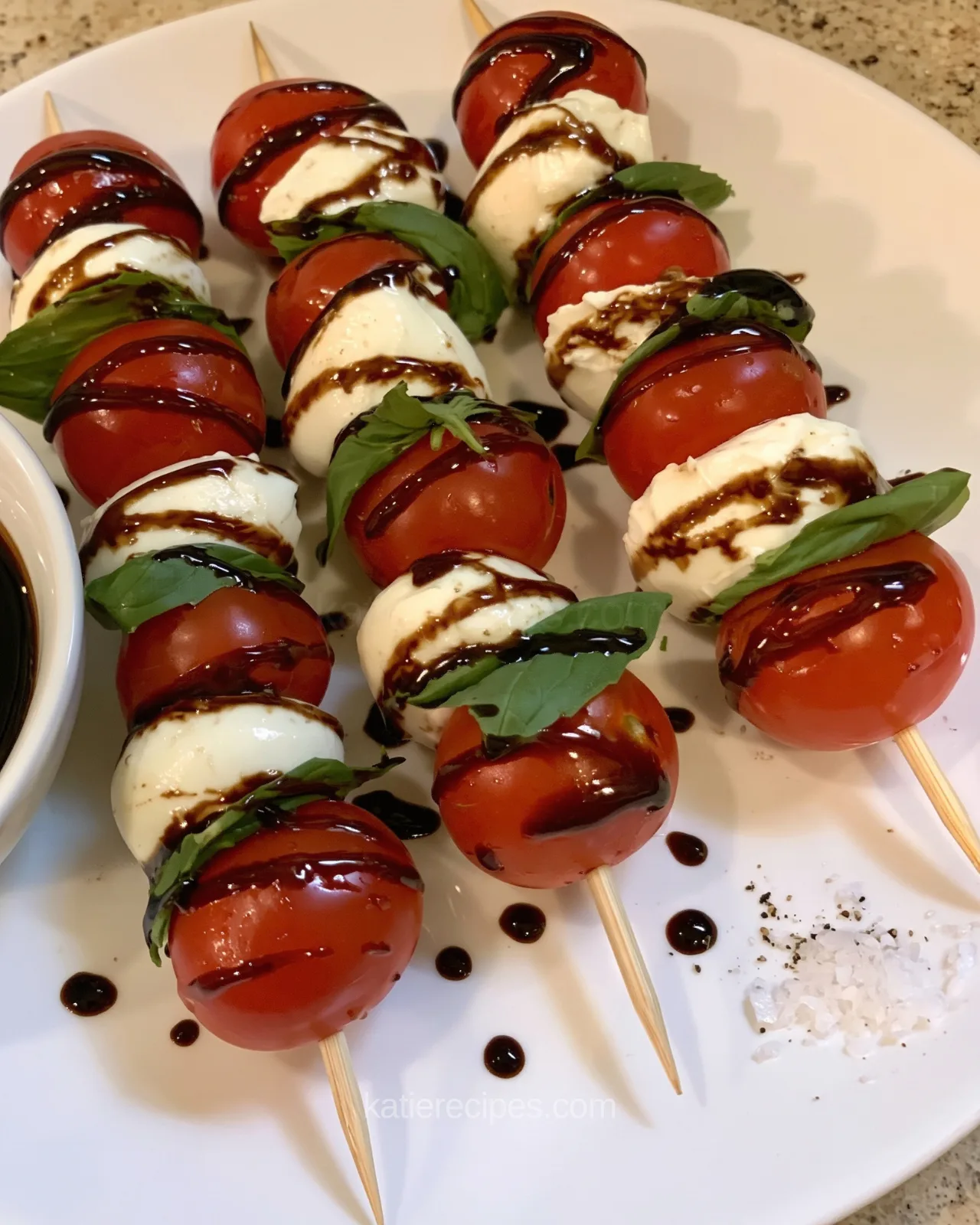Close-up of caprese skewers with balsamic glaze drizzled over tomatoes and mozzarella, with drops of glaze and flaky salt on a white plate