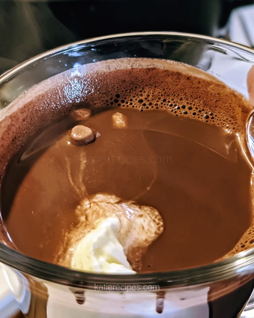 Close-up of a glass mug of hot chocolate with whipped cream and a foam ring at the rim, with a slow cooker lid behind