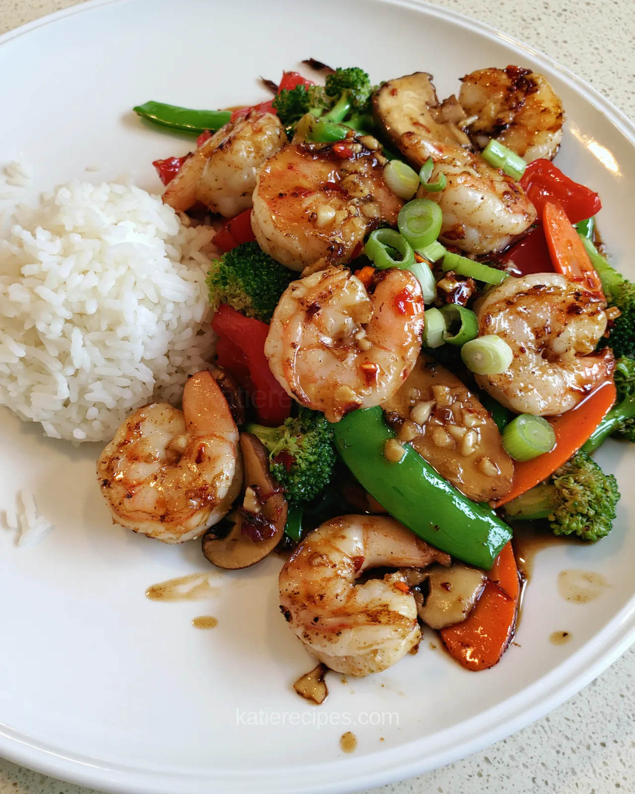 Close view of Hunan shrimp with seared edges, garlic and chili in a glossy sauce with broccoli, red pepper, carrot, mushrooms and snow peas, beside white rice