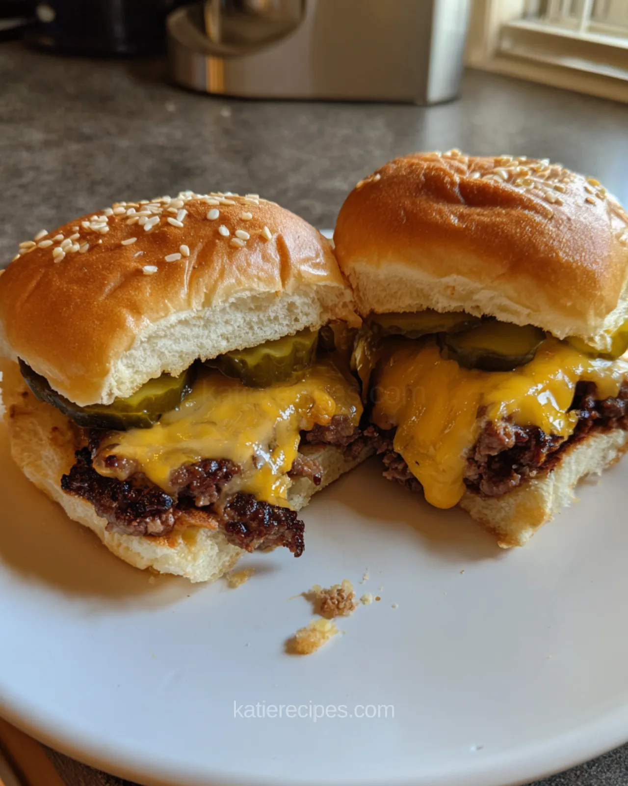 Hawaiian roll cheeseburger slider cut in half showing a browned beef patty, melted cheese, pickle chips and a golden sesame bun on a white plate
