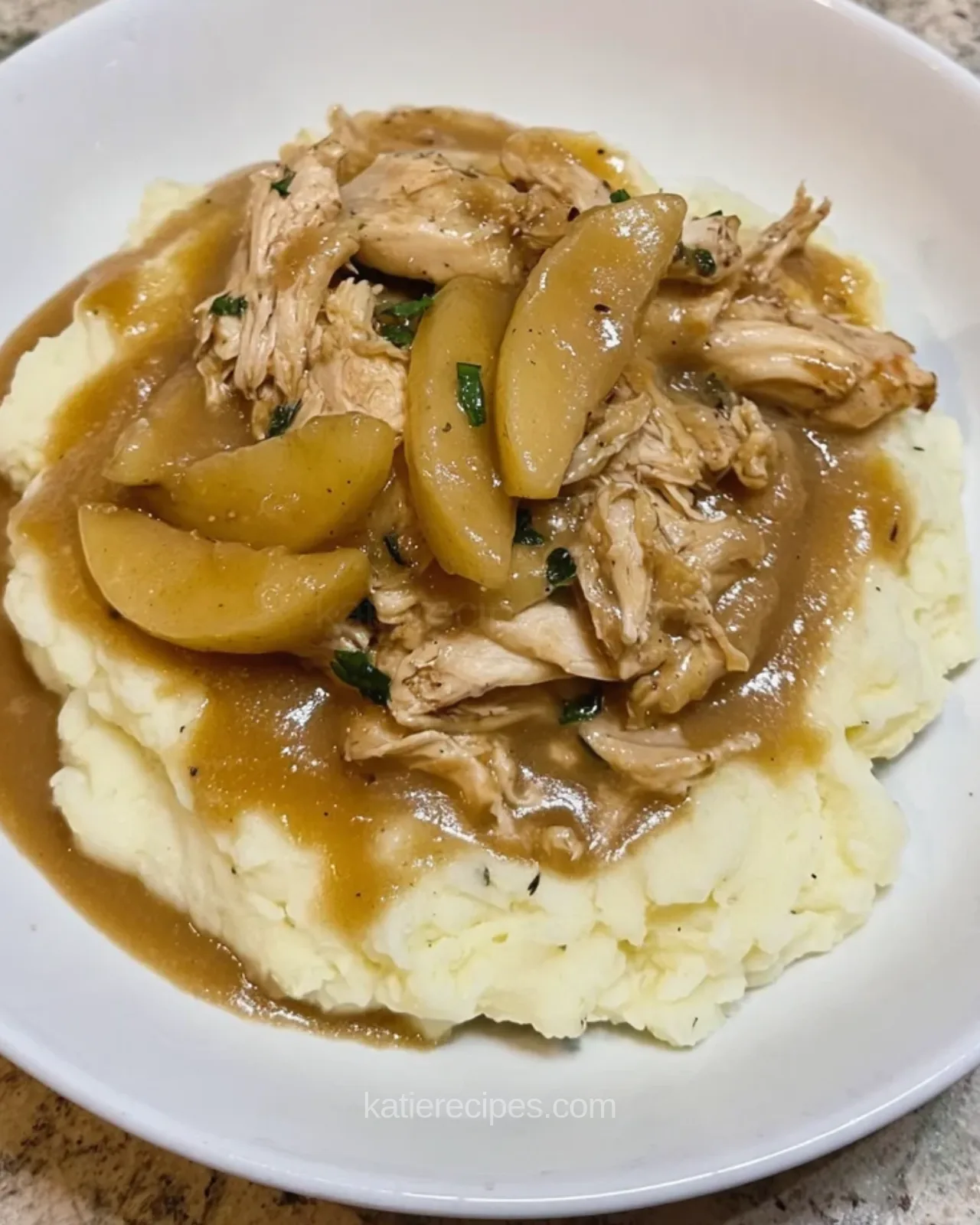 Shredded chicken and thick apple slices in a brown sauce over mashed potatoes in a white bowl, sprinkled with chopped green herbs