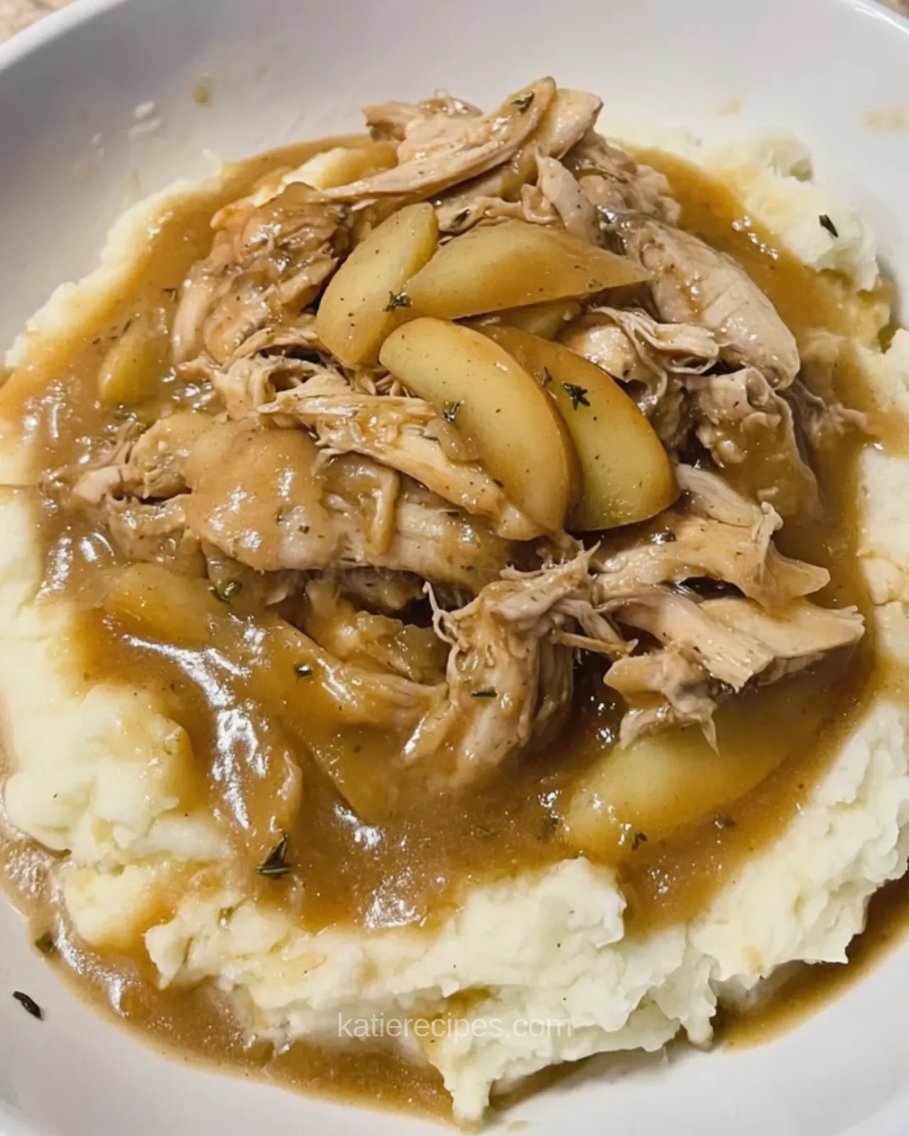 White bowl of mashed potatoes topped with shredded chicken, apple slices and brown sauce